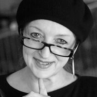 Schwarz-weiß Portraitaufnahme einer Frau mit Brille und Baskenmütze. Die Brille sitzt auf der unteren Nasenspitze, so dass sie über die Gläser hinweg in die Kamera schaut und offen lächelt. Ihren Zeigefinger hat sie an das Kinn gelegt.