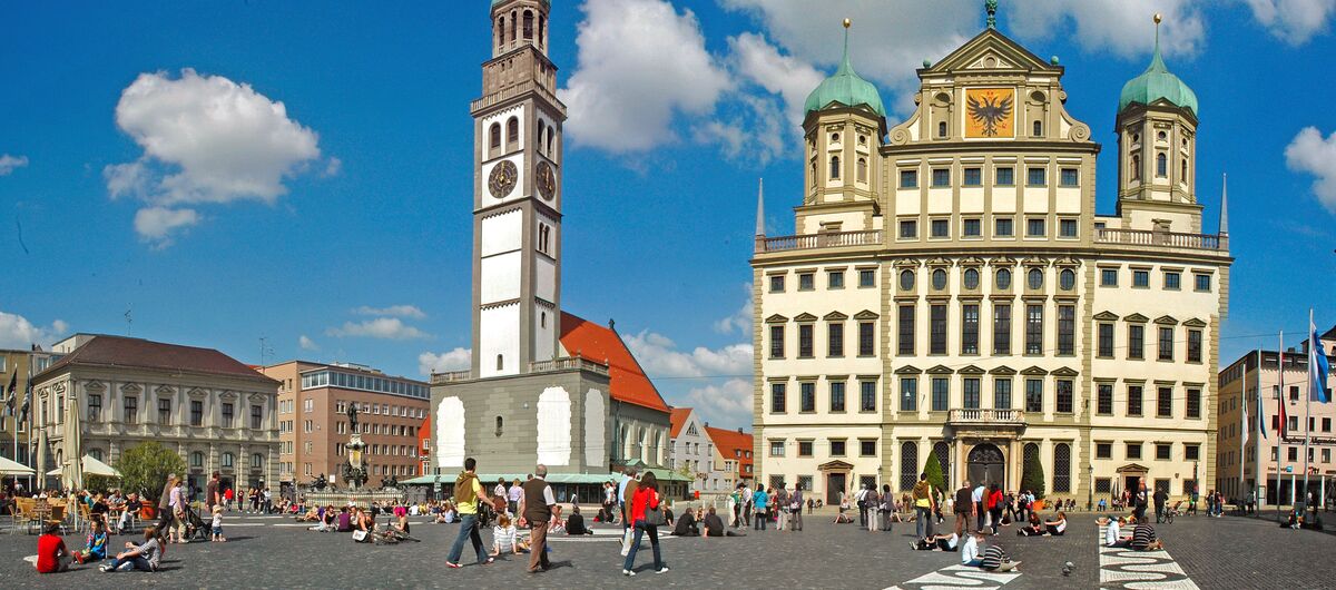 Foto des Marktplatzes von Augsburg. Im Hintergrund stechen eine Kirche mit hohem schmalen Glockenturm sowie das Augsburger Rathaus hervor. Der Marktplatz besteht aus dunklem Kopfsteinpflaster. Teils bilden weiße und dunkle Steine ein wellenartiges Muster.