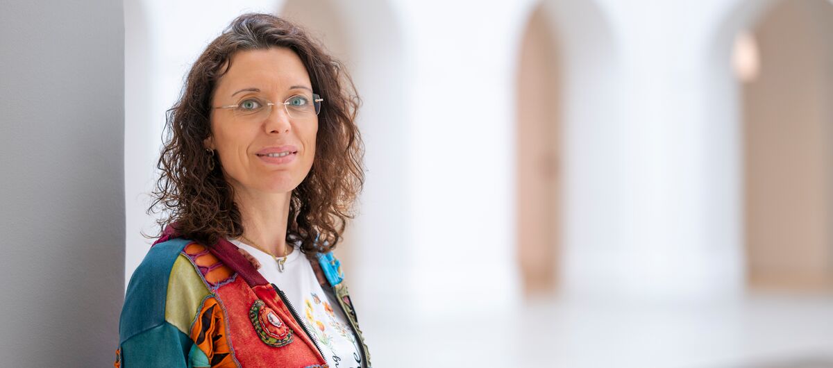 Foto in einem hellen schlichten Raum. Im Vordergrund ist auf der rechten Seite der Oberkörper eine Frau zu sehen. Sie schaut die Betrachtenden freundlich an. im Hintergrund sind Rundbogendurchgänge zu sehen.