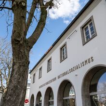 Foto der Außenansicht des Dokumentationszentrums Nationalsozialismus der Museen Freiburg. Fünf Rundbögen mit Fensterglas prägen die Hausfront im Erdgeschoss. Darüber ist der Schriftzug Dokumentationszentrum Nationalsozialismus zu lesen.