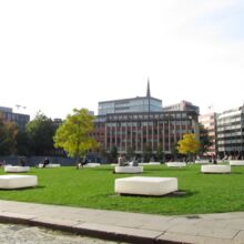 Weiße Leuchtkästen stehen auf grünem Rasen, vereinzelt sitzen Menschen auf ihnen. Die Fläche wird gesäumt von Bäumen und Bürogebäuden der Hamburger Innenstadt.