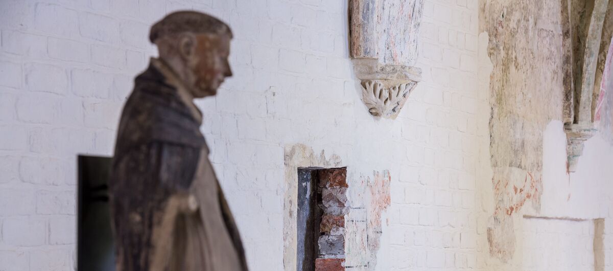 Foto in schräger Perspektive auf eine weiß gestrichene Backsteinwand., in die ein kleiner Durchbruch geschlagen wurde. Im Vordergrund steht eine Dominikanerstatue.