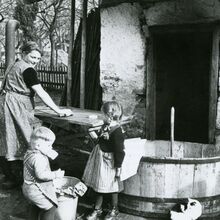 Schwarz-weiß Foto einer Alltagsszene einer Familie. Eine Frau arbeitet mit einem Mädchen und einem jüngeren Jungen vor einem Haus. Die Frau schaut freundlich über die Schulter zu den Kindern.