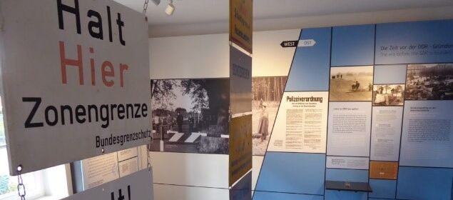 Foto eines Ausstellungsbereichs. Im Vordergrund auf der rechten Seite hängen Schilder untereinander in zwei Reihen, auf denen beispielweise steht: Halt Hier Zonengrenze Bundesgrenzschutz.