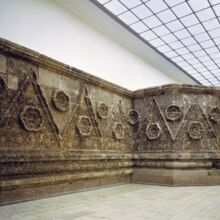 Hoher Raum mit originalgetreuer Mschatta-Fassade, die aus grauem Stein besteht, welcher mit hervorstehender Zickzack-Linie und steinernen Rosetten verziert ist.