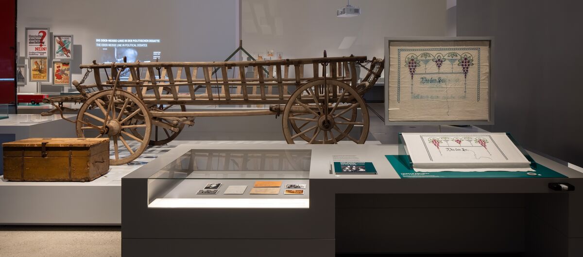 Foto in einen dunkel gehaltenen Teil des Ausstellungsraums. Hell erleuchtet sind Exponate, wie z. B. ein einfacher Kutschwagen aus Holz, eine Holztruhe, eine Vitrine mit Fotos und Unterlagen, eine verziertes weißes Stofftuch.