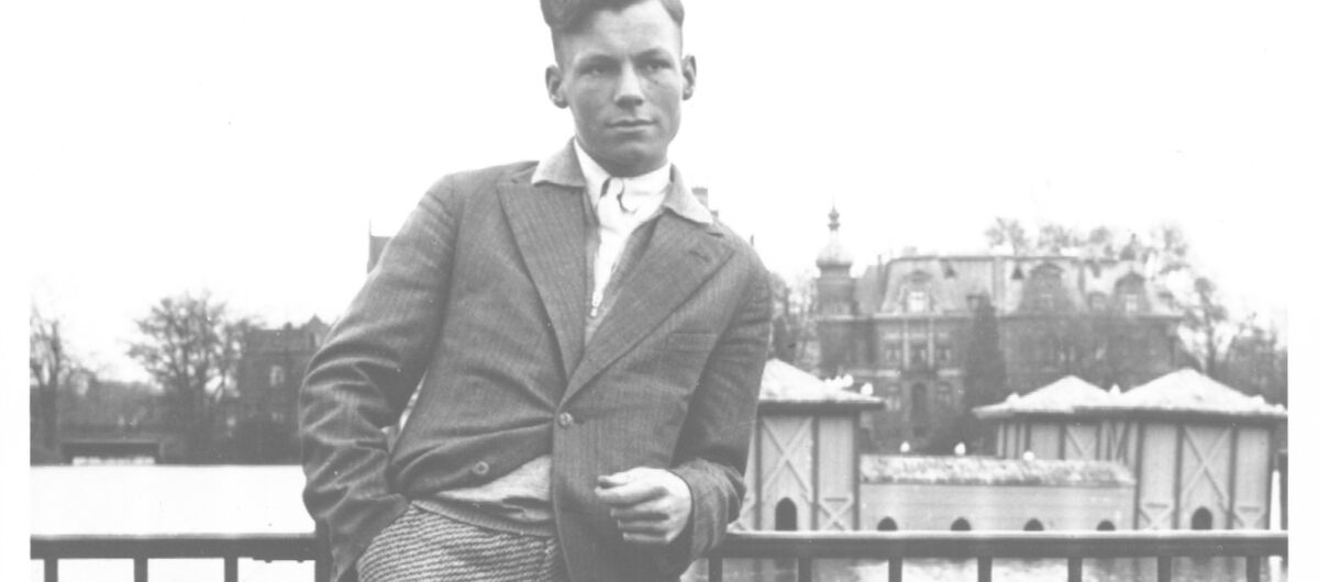 Schwarz-weiß Foto des jungen Willy Brandts in Jackett und Knickerbocker. Er steht vor einem Gewässer und lehnt sich mit seinem linken Arm auf einen Zaun. Die rechte Hand Steckt in seiner Hosentasche. Er schaut mit neutralem Gesichtsausdruck nach links.