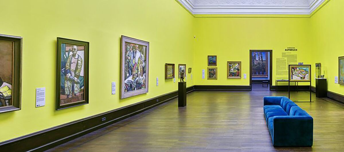 Gelb gestrichener Ausstellungsraum mit Oberlicht, blauem Sofa und unterschiedlich großen, gerahmten Bildern an der Wand