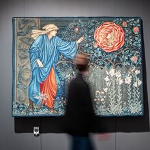 Foto eines angeleuchteten Teppichs vor einer grauen Wand, vor dem ein verschwommener Mensch herläuft. Auf dem Teppich ist ein Mann im Garten abgebildet. Er streckt seinen linken Arm zu einer überdimensionierten Rose mit Frauenkopf.