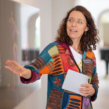 Foto eines Ausstellungsbereichs. Im Vordergrund ist eine Frau mit Zettel und Stift in ihrer linken Hand zu sehen, die spricht und mit ihrer rechten Hand auf ein großes Foto rechts neben ihr zeigt. Ihre Augen sind auf das Foto gerichtet.