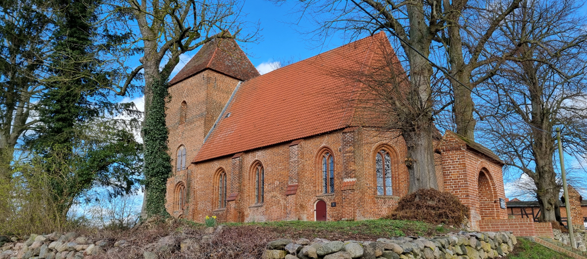 Foto der Außenansicht der Dorfkirche Friedrichshagen, die leicht erhöht steht und von einer Natursteinmauer umgebene ist. Die Kirche besteht aus roten Ziegeln und hat ein rotes Dach. Drumherum stehen hohe unbelaubte Bäume.