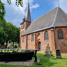 Foto der Fischerkirche St. Andreas in Lübeck. Die Backsteinkirche ist umgeben von Rasenfläche, auf denen historische Grabsteine stehen, und hohen Bäumen. Im Vordergrund der Kirche steht auf einer Fläche mit kleinen Kieselsteinen ein schwarzes Fischerboot.