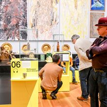 Ausstellungsansicht zu "Hör mal hin!". Besuchende - teils mit Kopfhörern auf dem Ohr - schauen sich Objekte in Vitrinen und auf Podesten sowie Gemälde an Wänden an. Die Objekte sind teils religiöser Herkunft, die Gemälde abstrakt bis realistisch.