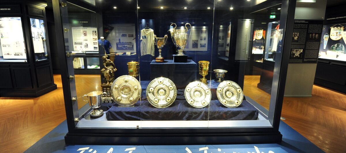 Vitrine mit Pokalen und Meisterschalen des HSV. Davor ein Schriftzug mit "Titel, Tore und Triumphe".  Im Hintergrund Trikots. und weitere Ausstellungsvitrinen.