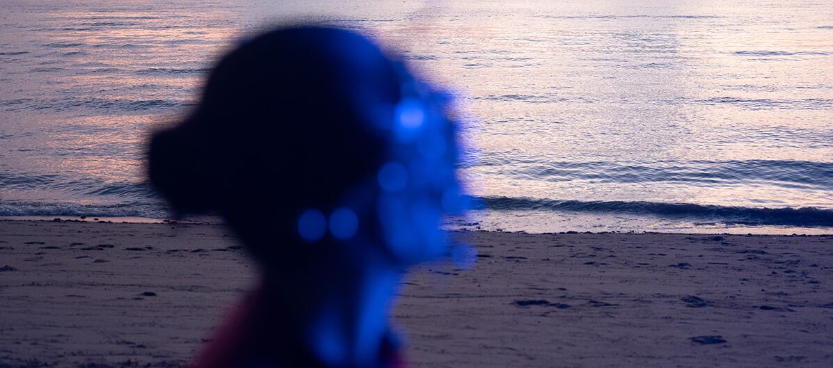 Foto, das im Vordergrund einen blau angeleuchteten Oberkörper einer Person zeigt. Er ist verschwommen. Der Hintergrund ist scharf. Zu sehen ist in dämmeriger Stimmung ein Strand. Das Meer ist ruhig. Wolken sind am Himmel.