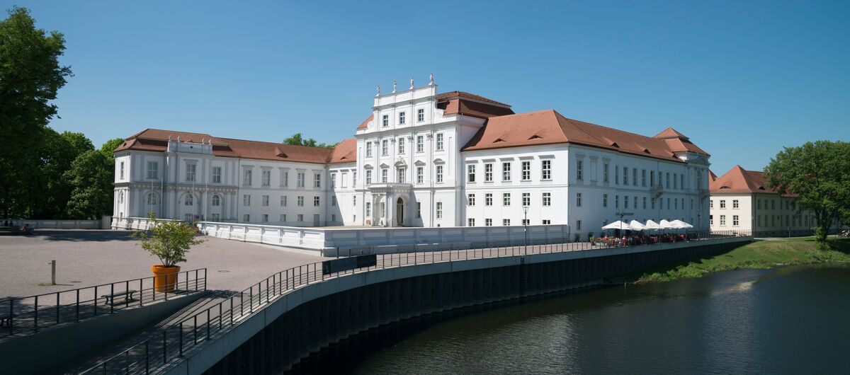 Foto des Schloss Oranienburg. Ein prächtiges weißes Gebäude mit rotem Dach steht im Zentrum des Fotos. Die rechte untere Ecke dominiert ein ruhiges Gewässer. Das Wetter ist sonnig.