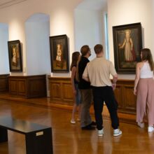 Blick in Ausstellungsraum mit Boden und hüfthohen Wandvertäfelungen aus Holz. Vier Gemälde hängen an den Wänden. Um das ganz rechts im Bild stehen zwei Frauen und zwei Männer und betrachten es. Links unten steht eine schwarze minimalistische Besucherbank.