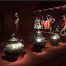 Foto von drei metallischen Gefäßen unterschiedlicher Form. Sie stehen auf einer dunkelroten Oberfläche. Der Hintergrund ist sehr dunkel. Angeleuchtet stehen dort eine Statur sowie ein Bild, das eine Szene mit drei Menschen vergangener Zeiten darstellt.