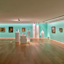Foto eines Ausstellungsbereichs. An einer helltürkiesen Wand hängen in einer Reihe verschiedenste alte Gemälde in Rahmen. Davor stehen vier schmale Vitrinen, in denen sich Gefäße befinden.