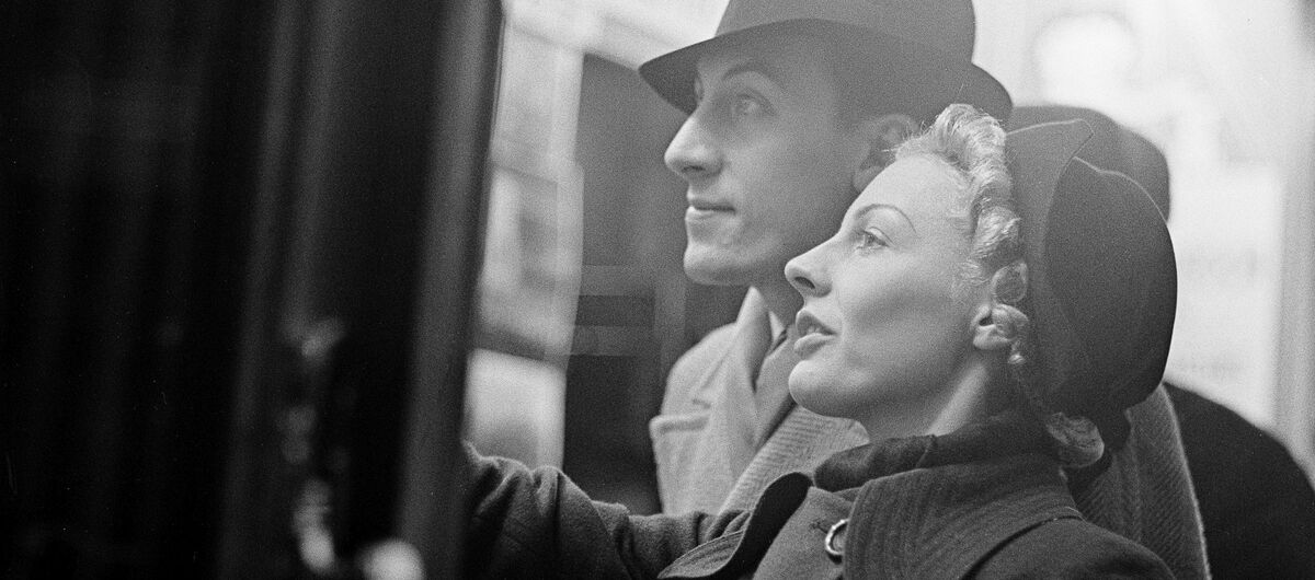 Schwarz-weiß Foto einer Frau und eines Mannes, die Ihren Blick auf etwas an der linken Fotoseite richten. Der rechte Arm der Frau ist leicht gehoben, alsob sie auf etwas zeigt. Beide sind mit Hut und Mantel fein gekleidet.