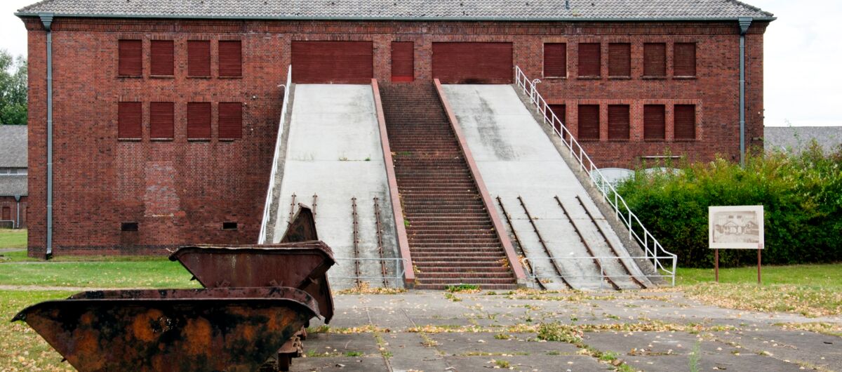 Foto von einem Klinkergebäude in das zwei Straßen hochführen. Dazwischen sind Treppenstufen angelegt. Beide Straßen haben je zwei Schienensysteme für Loren. Die Fenster und Tore sind mit Brettern verschlossen. Im Vordergrund stehen zwei verrostete Loren.