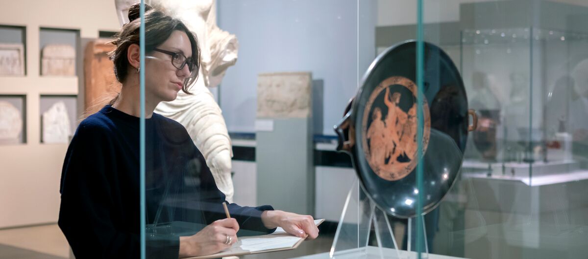 Foto eines Ausstellungsbereichs. Vor einer Glasvitrine, in der eine antike Schale ausgestellt ist, steht eine Frau mit Stift in ihrer rechten Hand, die auf einem dünnen Brett mit Papier liegt. Sie betrachtet das ausgestellte Objekt.