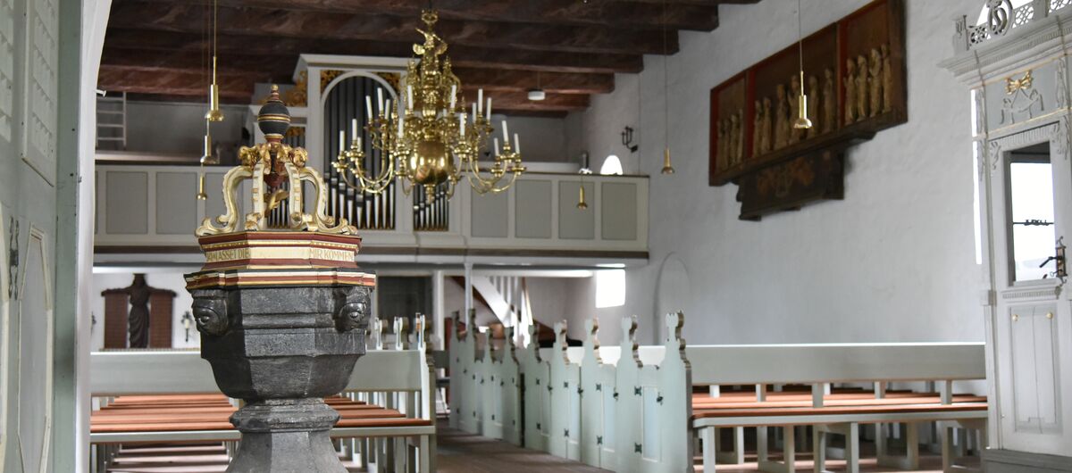Foto des Kirchenraums. Im Vordergrund ist das Taufbecken zu sehen. Dahinter sind die Kirchbänke in zwei Reihen nebeneinander aufgestellt - oberhalb dazwischen ein Kronleuchter. Im Hintergrund ist die Orgel zu sehen. Diese Decke besteht aus Holzbalken.