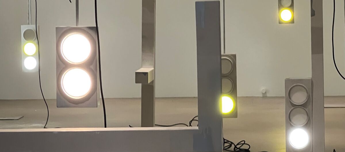 Foto einer Installationsansicht. An fünf auf dem Boden stehenden Ständern hängen fünf ampelartige Lichter, die weiß bis gelb leuchten. Ein Licht hat zwei statt drei Lichtkreise. Es leuchten insgesamt sieben von 14 Lichtkreisen. Kabel liegen herum.