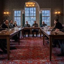 Filmausschnitt aus dem Film "Die Wannsee-Konferenz". An u-förmig gestellten Holztischen in einem Raum mit großen Fenstern und verzierten Wänden sitzen 13 Männer und zwei Frauen. Alle tragen Anzug. Es liegen Papiere und Mappen auf den Tischen.
