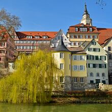 Foto des Hölderlinturms von Seiten des Fluss Neckar. Hinter dem runden Turm mit spitzem Dach schließt sich ein Gebäude an. Die unterste Etage des Gebäudekomplexes ist aus Naturstein, die Fassade der oberen beiden Etagen gelb gestrichen.