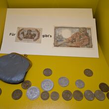 Foto von 18 Münzen unterschiedlicher Größe und Farbe. Links daneben liegt ein kleines graues Münzportemonnaie. Darüber liegt ein Papier auf dem ein kleiner und ein großer Geldschein kleben. Es steht: Für "kleiner Geldschein" gibt`s "großer Geldschein".