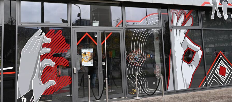 Foto des Eingangsbereichs zum Illuseum in Berlin. Die Glasfront ist mit großen Aufklebern beklebt, die z. B. abstrakte Hände abbilden, die einen Schatten in Form eines Bären werfen.