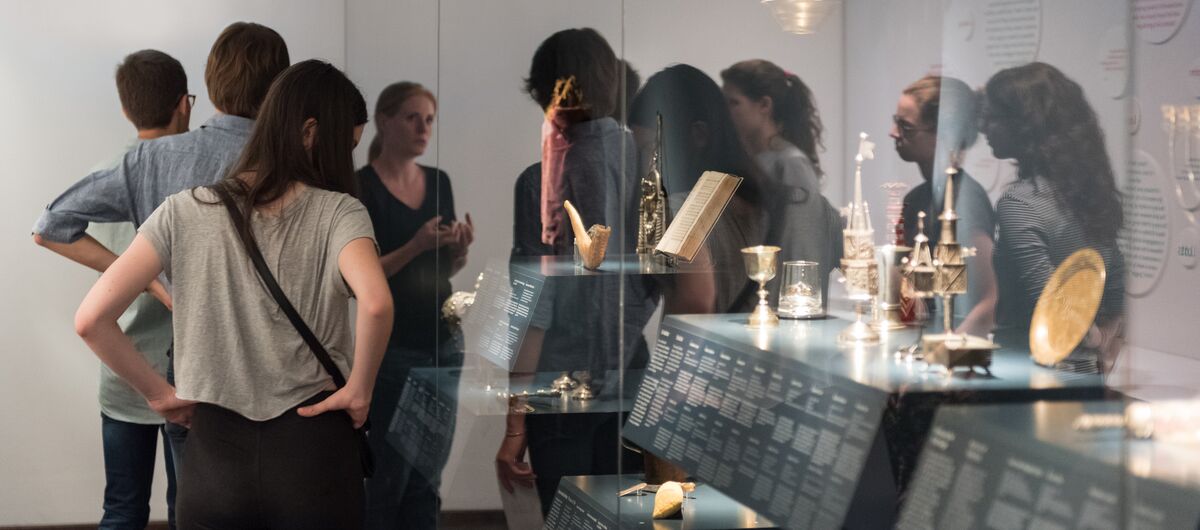 Foto eines Ausstellungsbereichs. Um eine große Vitrine mit metallischen Gegenständen und einem Buch ist eine Gruppe versammelt, die eine Führung erhält.