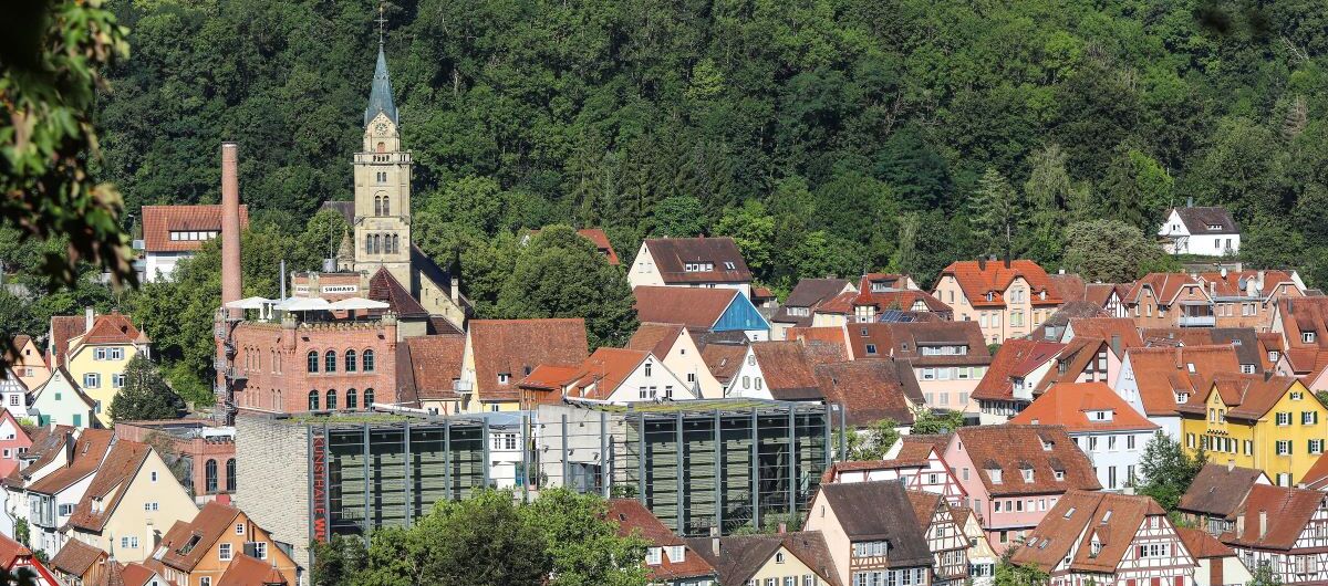 Foto von der Stadt Schwäbisch Hall. Im Hintergrund steht ein Wald. Die Kunsthalle Würth steht im Zentrum des Fotos. Sie besticht durch eine Gläserne Front. Drumherum stehen Fachwerkhäuser, dahinter eine Kirche mit Turm.