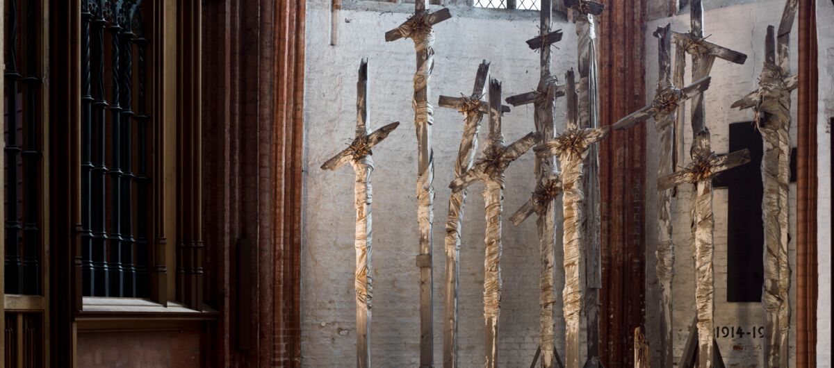 Blick in den Innenteil einer Kirche, die links eine Art Kanzel oder Beichtstuhl zeigt und rechts eine Ansammlung mehrerer Holzkreuze. Sie sind fast so hoch wie die Hälfte des Raumes, überragen die Mauer, in denen die Fenster sitzen.