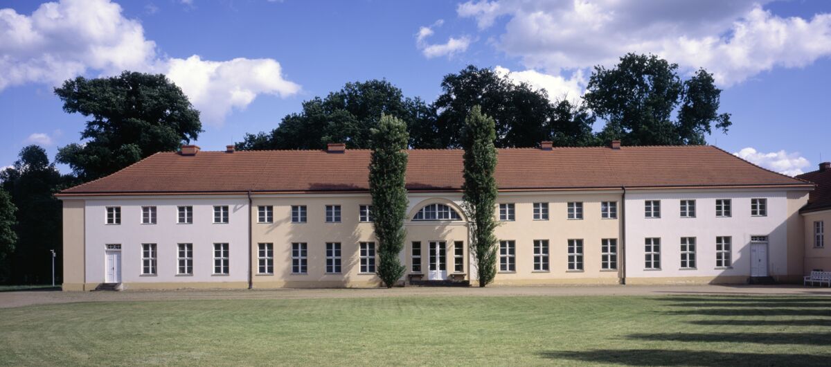 Foto vom Schloss Paretz. Davor eine große Rasenfläche. Der Eingang des Gebäudes wird durch zwei hohe schmale Bäume gesäumt. Das Gebäude ist schlicht gehalten. Der Mittelteil ist in einem Vanilleton gestrichen, die beiden äußeren Teile in weiß.