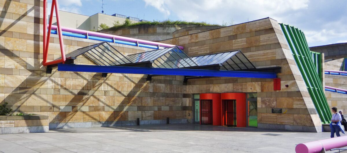 Außenansicht des Eingangsbereichs der Staatsgalerie Stuttgart. Das Sandsteingebäude wird durch moderne bunte Stahlelemente ergänzt. Die beiden zylinderförmigen Eingangselemente sind rot. Eine gebogene Fensterfront ist durch grüne Stahlträger unterbrochen.