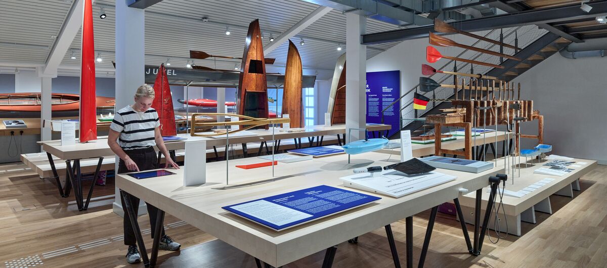 Foto eines Ausstellungsbereichs des Wassersportmuseums. Auf großen Tischen sind Informationsschilder, Paddel und Kanus ausgestellt.