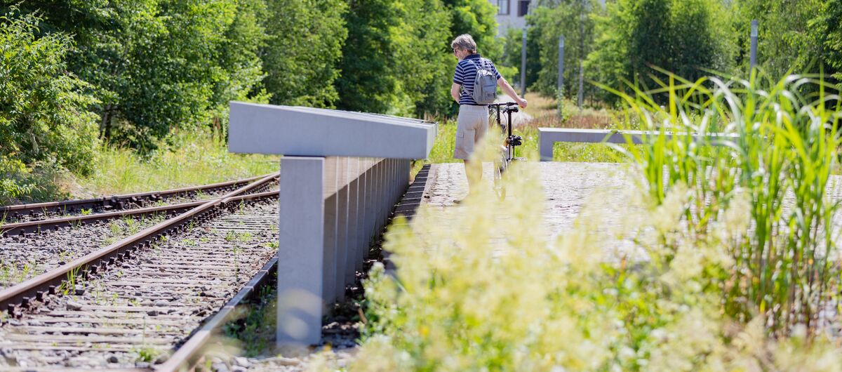 Foto mit Blick aus Kniehöhe. Links sind zwei Gleise zu sehen. Mittig eine lange Reihe von Informationstafeln aus Beton, die von einem Menschen mit Fahrrade betrachtet werden. Im Hintergrund sind grüne Bäume sowie eine Reihenhauszeile zu sehen.