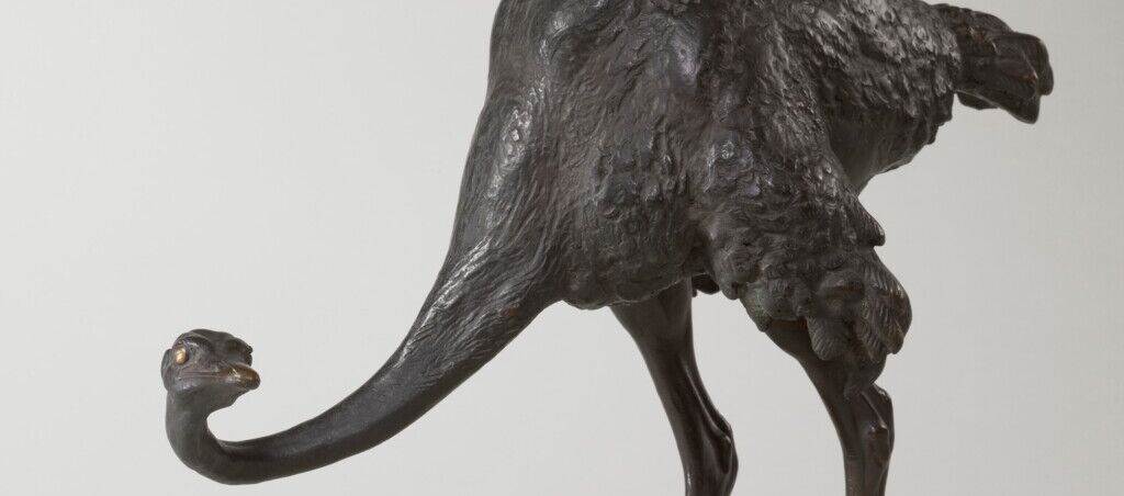Foto eines Straußes aus Bronze. Dieser steht auf einem runden Steinsockel. Der Strauß steht mit leicht vorgebeugtem Hals und schaut vom Betrachtenden aus gesehen nach rechts.
