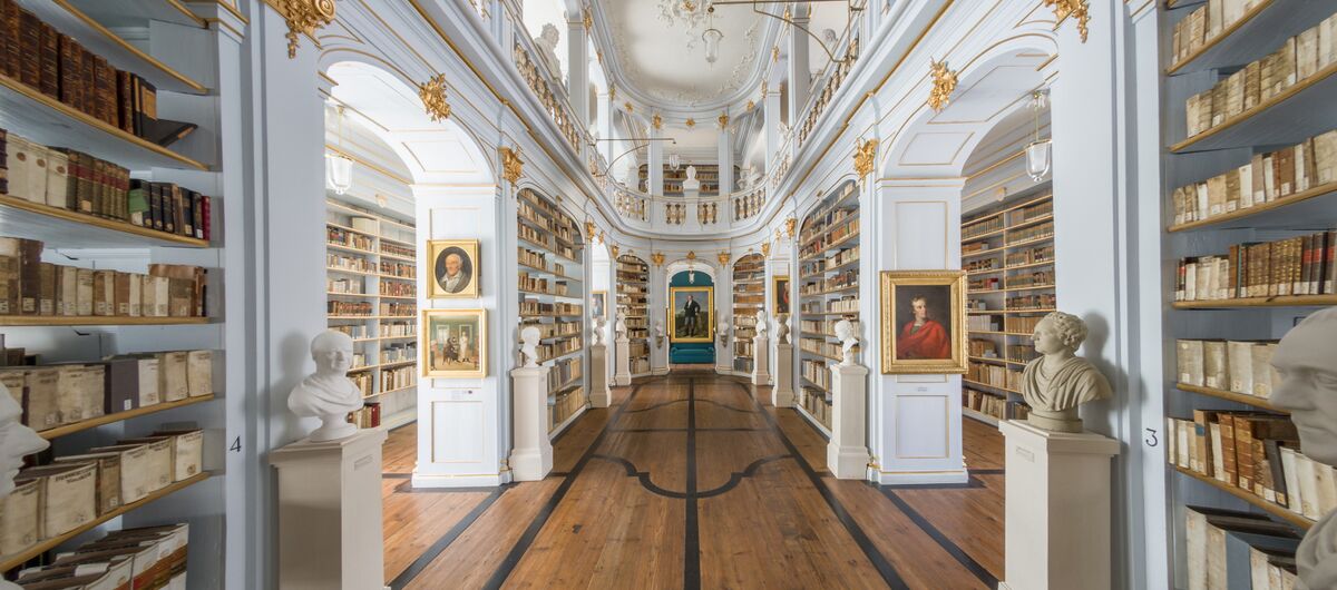 Foto vom historischen Bibliothekssaal mit breitem Holzgang unter kunstvoll verzierten Deckengewölbe. Die Wände zieren Regale mit goldenen Elementen, die Bücher, Porträtgemälden und Büsten enthalten.