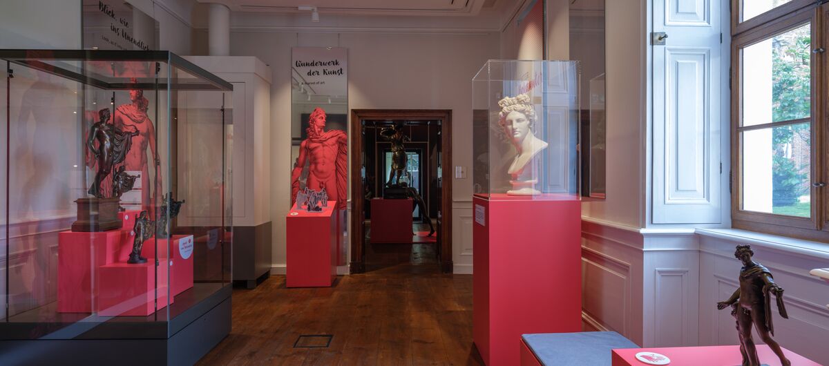 Foto eines Ausstellungsbereichs. In dem Raum mit großen Fenster auf der rechten Seite stehen auf dunklem Holzboden Vitrinen mit roten Sockel. In diesen ist eine weiße Büste und und dunkle Figuren zusehen.