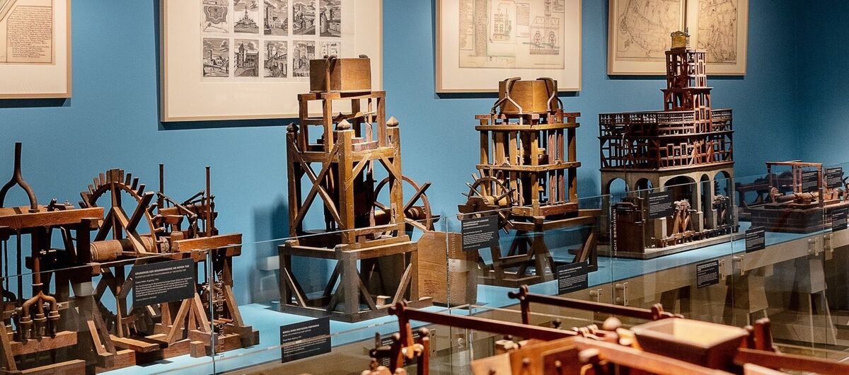 Foto der Modellkammer des Maximilianmuseums. Zu sehen sind fünf verschiedene Maschinen, eine weitere steht im Vordergrund. An der Wand dahinter hängen vier Bilder mit Skizzen und Karten.