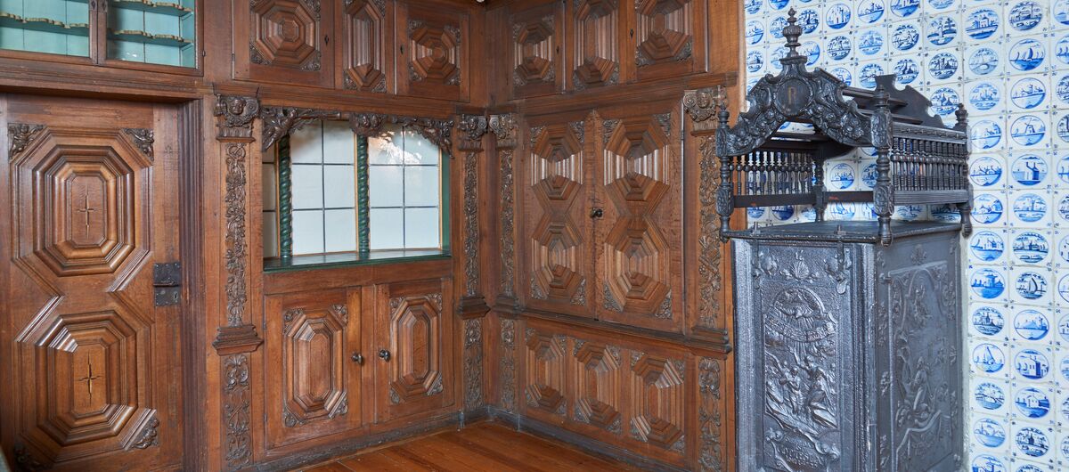 Foto einer barocken Bauernstube. Der Boden ist ebenso wie die kunstvoll geschnitzte Tür und die Wände links und mittig im Foto aus dunklem Holz. Die rechte Fotoseite wird durch eine mit blau-weißen kleinen Fliesen und einem kunstvollen Metallofen geprägt.