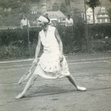 Schwarz-weiß Foto einer Frau in weißem Kleid und mit weißem Haarband mit Tennisschläger in ihrer rechten Hand. Sie steht im Ausfallschritt auf einem Tennisplatz und lächelt. Im Hintergrund stehen zwei Männer und schauen zu.