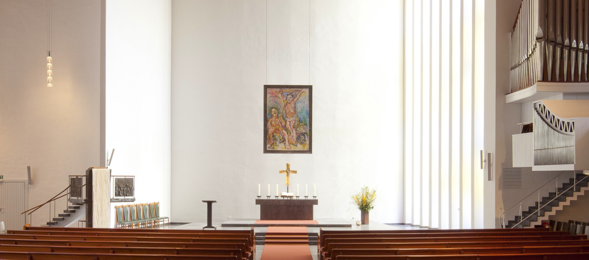 Innenraum einer modernen Kirche. Weiße hohe Wände, ein fast ebenerdiger Altarbereich, der über drei Stufen erhöht steht. Ein goldenes Kreuz und Kerzen stehen auf ihm. Links eine kleine steinerne Kanzel, rechts die Orgel. Die Sitzbänke sind aus Holz.