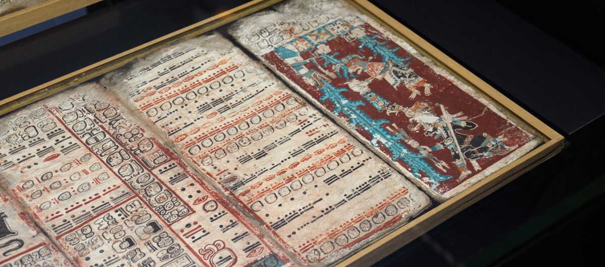 Fot einer Maya-Handschrift. Auf einem schwarzen Untergrund liegt in einem hellen dünnen Holzrahmen mit verschiedenen Faarben beschrieben und bemalte Papiere. Die Ecken der Seiten sind abgenutzt.