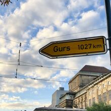 Foto eines gelben Straßenschildes, auf dem Gurs 1027 km steht. Es zeigt nach links. Im Hintergrund sind Teile von älteren und neueren Gebäuden sowie Oberleitungen von Straßenbahnen zu sehen.