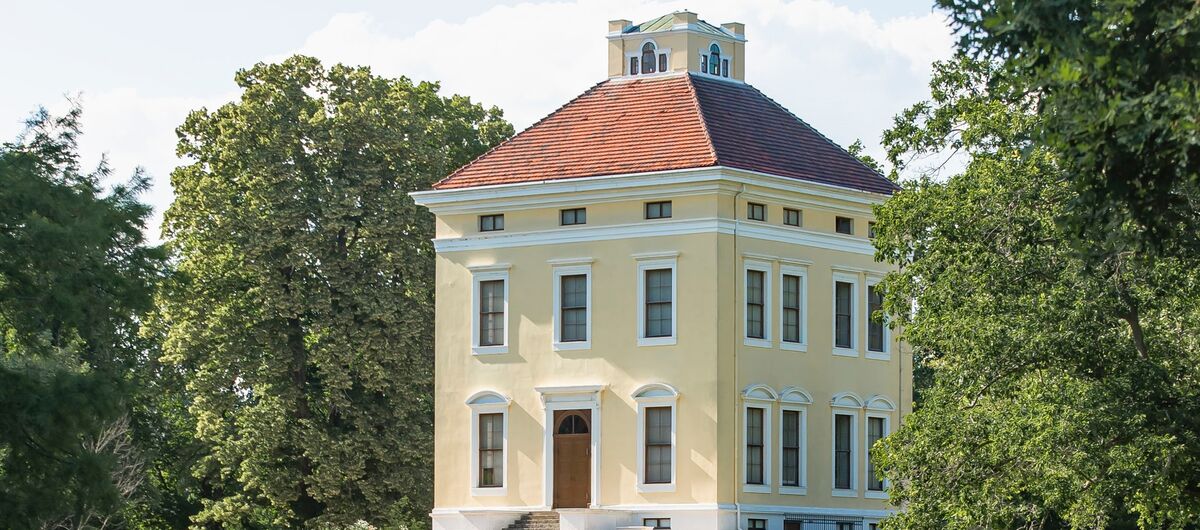 Foto des Schloss Luisium. Es ist ein historischer würfelartiger dreistöckiger Bau mit rotem Ziegeldach in einem Park. Auf dem Dach ist ein flaches Türmchen mit Fenstern draufgesetzt. Das Schloss ist hellgelb gestrichen, die Fenster weiß umrandet.