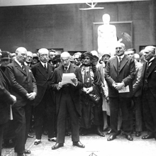 Schwarz-weiß Foto einer Gruppe von Männern und Frauen in einem Raum, die sich um einen Mann versammeln, der ein Blattpapier in den Händen hält und etwas vorzulesen scheint. Alle sind schick gekleidet und haben eine neutrale bis ernste Miene.